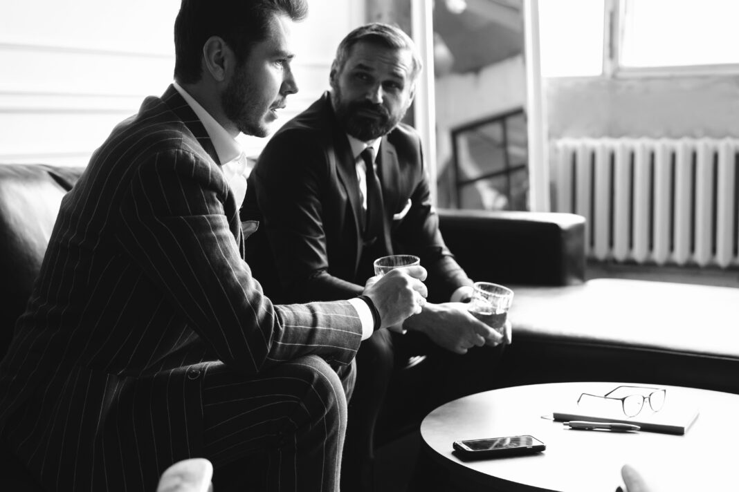 Relaxing. Full length of two young handsome men in suits holding glasses and looking at each other while resting indoors.